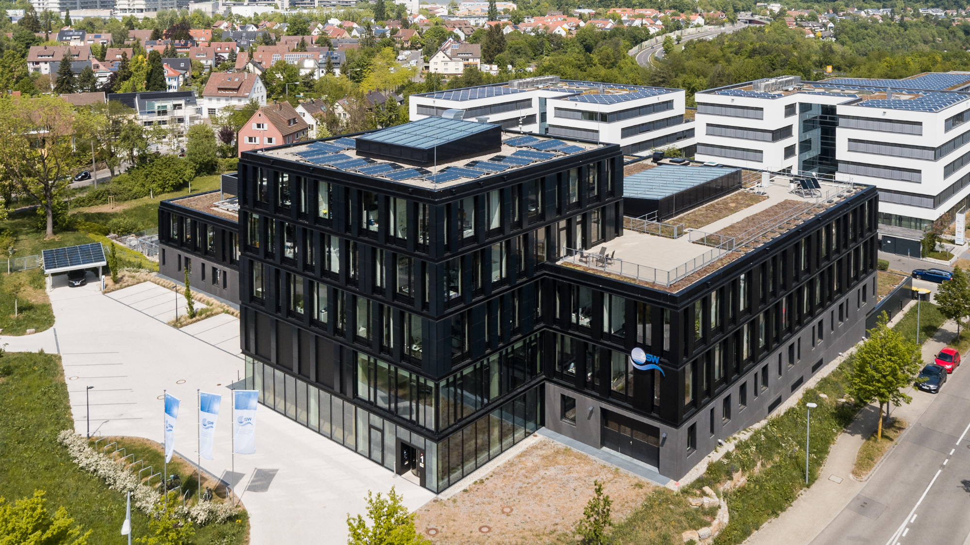 Luftaufnahme des ZSW-Gebäudes in Stuttgart-Vaihingen. Im Vordergrund ist der fünfstöckige sogenannte Turm mit einer beinahe quadratischen Grundfläche zu sehen. Hier befindet sich der Besuchereingang zum Institut. Nach hinten versetzt wird der Turm vom niedrigeren Rest des Institutsgebäudes auf drei Seiten umfasst. Das Gebäude hebt sich durch die schwarze Fassade deutlich von den Gebäuden im Hintergrund ab. 